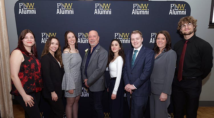 People in dress clothes pose in front of a PNW Alumni banner