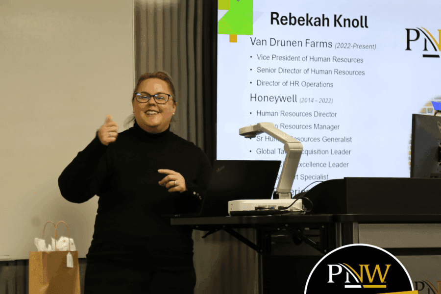 A smiling woman dressed in black giving a presentation next to a lectern with her name and work history in the background