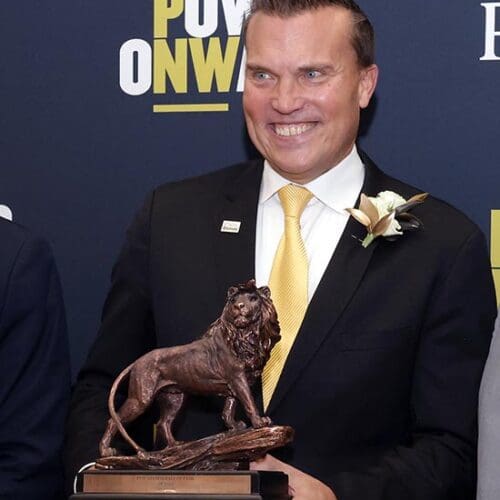 Jeff Strack in a suit and tie poses with his PNW Alumni Hall of Fame award (a lion statuette).