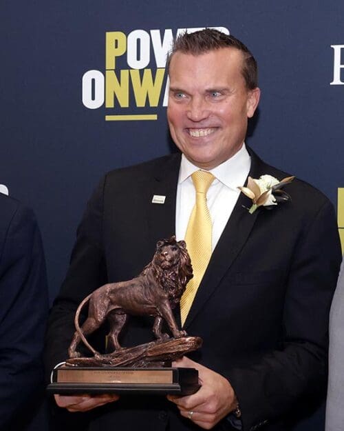 Jeff Strack in a suit and tie poses with his PNW Alumni Hall of Fame award (a lion statuette).