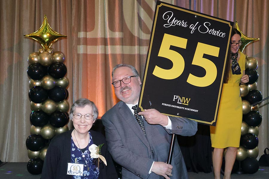 A woman and man pose at an event, with balloons in the background. The man is holding a sign that says Years of Service 55 PNW.
