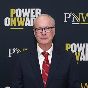 Brian Miller in a suit and tie before a PNW/Power Onward banner.