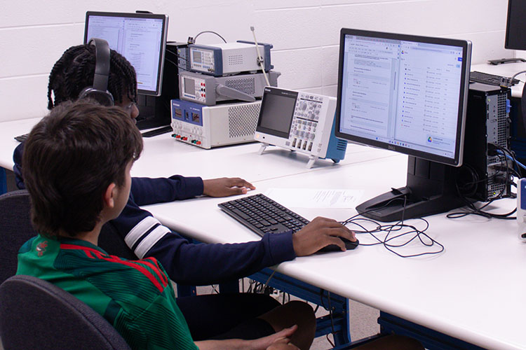 Two students sit at a computer and work together. One student has the keyboard and mouse in front of them, the other is watching.