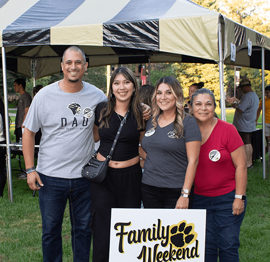A family of four poses together behind a yard sign that says "Family Weekend".