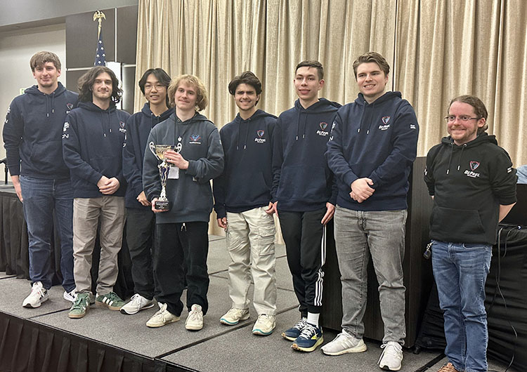 A group of students stands together on stage. They are wearing matching dark blue sweatshirts.