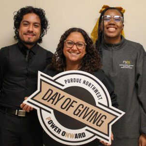 Three students pose together. The middle student is holding a PNW Day of Giving sign.
