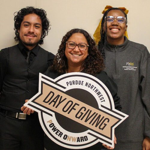 Three students pose together. The middle student is holding a PNW Day of Giving sign.