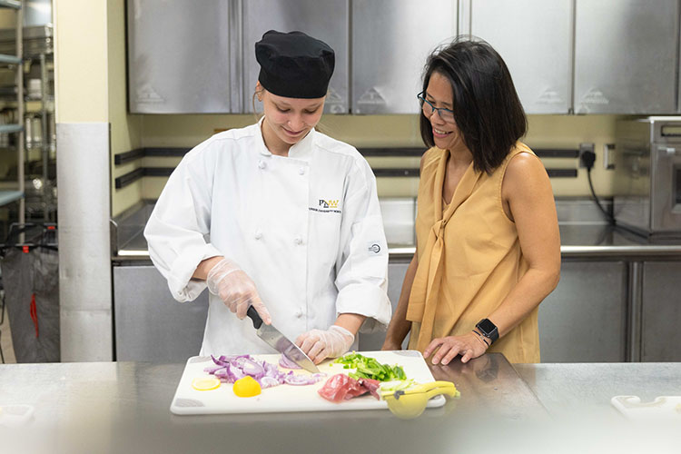 PNW student cutting up vegetables in the kitchen at the HTM building. Their professor is standing next to them.