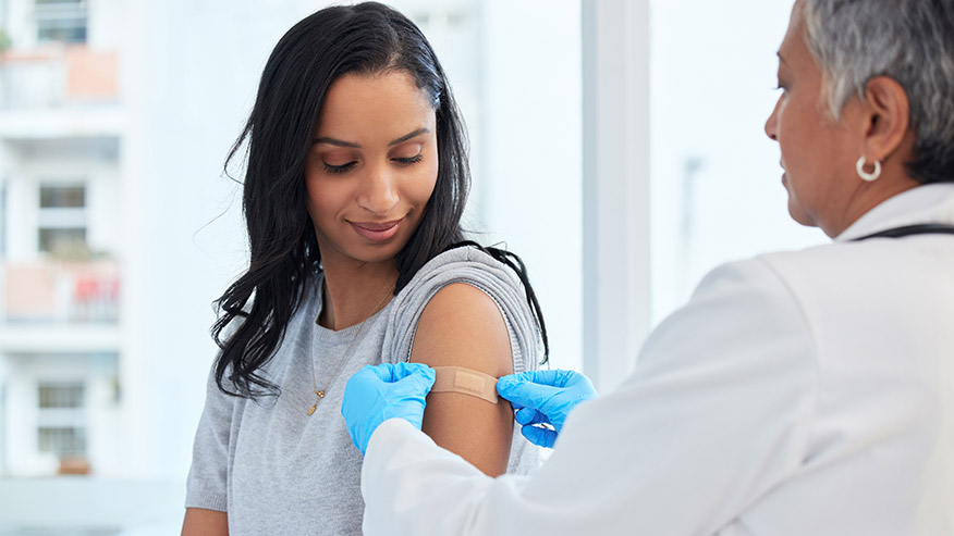 Healthcare professional wearing blue gloves places a bandage on a patient’s upper arm after an injection during a medical appointment in a bright clinical setting.