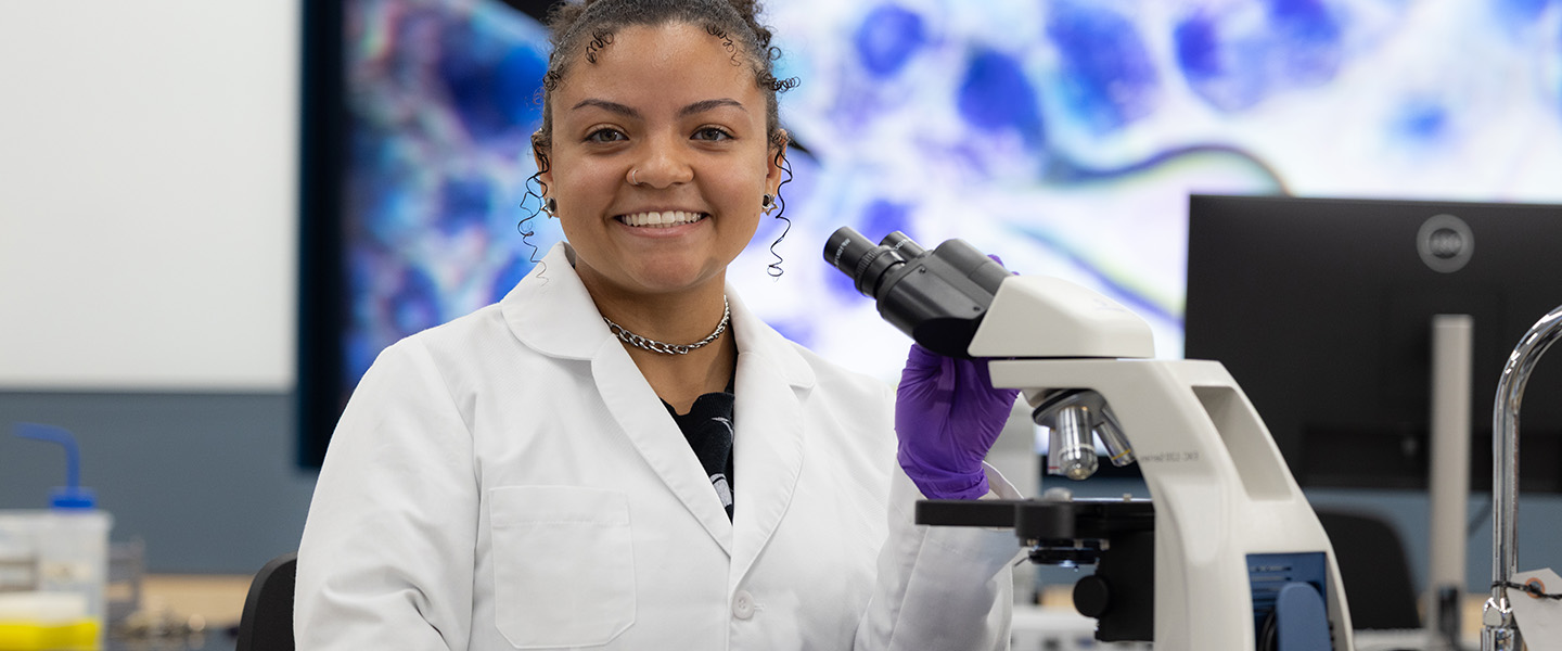 A student in a white lab coat sits at a microscope.