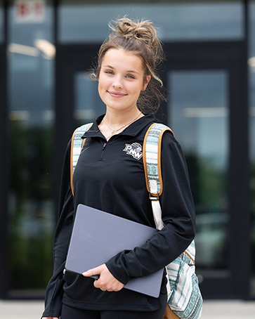 A student stands outside, wearing a backpack and holding a laptop.