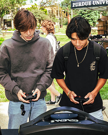 Two students stand outdoors and play on a game console.