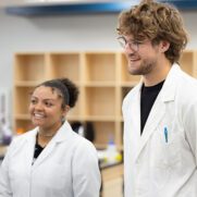 Two students stand in lab coats inside a lab classroom.