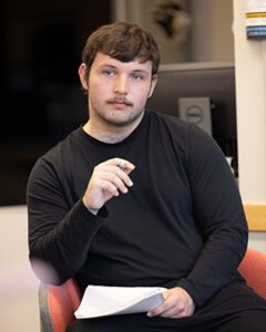 A student sits in a chair. They are holding a pencil in one hand and papers in the other.