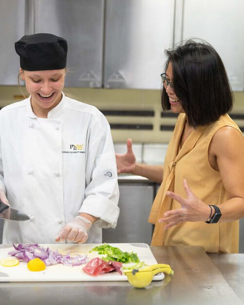 A student stands with an HTM instructor in the kitchen. The student is chopping food on a chopping board.