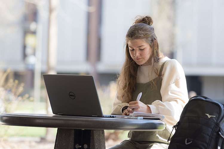 A student sits outside and writes in a notebook. They have an open laptop on the table in front of them.